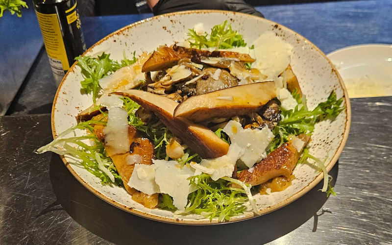 A restaurant plate with a bed of greens, shavings of cheese and sauteed nebrodini bianco mushrooms.